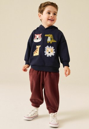 Young boy smiling wearing a navy hoodie with animal faces and loose red pants with white sneakers, standing on a plain light background.