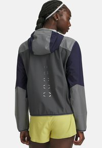 Water-resistant jacket in gray with navy accents, featuring reflective details, a hood, and a smooth texture. Paired with yellow shorts.
