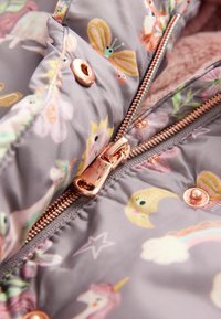 Grau gesteppte Jacke mit floralen und verspielten Mustern, ausgestattet mit einem roségoldenen Reißverschluss und Druckknöpfen, weiche Textilfutter sichtbar.
