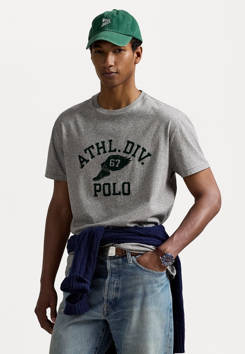 Grijze katoenen T-shirt met de opdruk "ATHL.DIV. 67 POLO" in donkergroen. Draag het met blauwe spijkerbroek en een marineblauwe trui om de taille geknoopt. Groene pet.