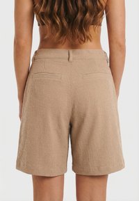 Mujer con cabello largo, vistiendo pantalones cortos beige de talle alto y textura, vista desde atrás, de pie contra un fondo claro y sencillo.