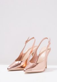 Escarpins slingback métalliques en or rose avec un design à bout pointu, des fines brides et un haut talon aiguille. Texture lisse, accents minimaux.