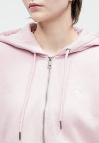 Person wearing a light pink hoodie with a silver zipper and drawstrings, featuring a small embroidered moose logo on the chest.
