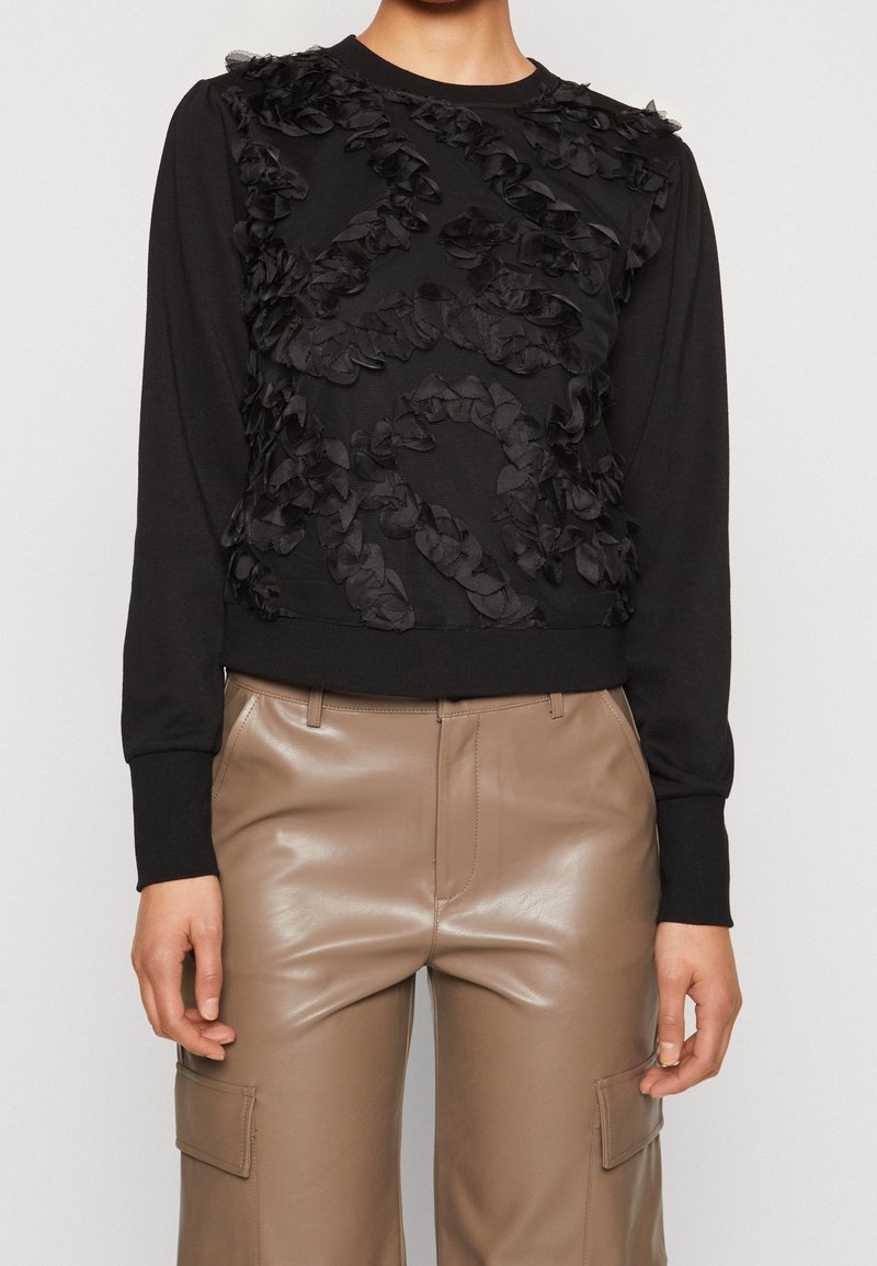 Femme portant un haut noir à manches longues avec des détails en volants texturés et un pantalon en cuir beige taille haute avec poches cargo.