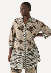 Beige-pitsi blouse, jossa on musta-valkoinen raidallinen kaulus, pitkät hihat ja teksturoitu musta-valkoinen kuvioitu helma.