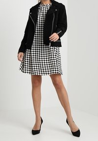 Svart mocka bikerjacka över en vit klänning med stora svarta prickar. Klänningen har en utställd form och kombineras med svarta klackar med spetsig tå.