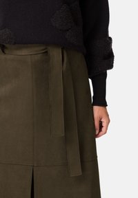 Spódnica z oliwkowego zamszu o gładkiej fakturze, z przednim rozcięciem i pasującym paskiem do wiązania. Widoczna górna część to czarny sweter.