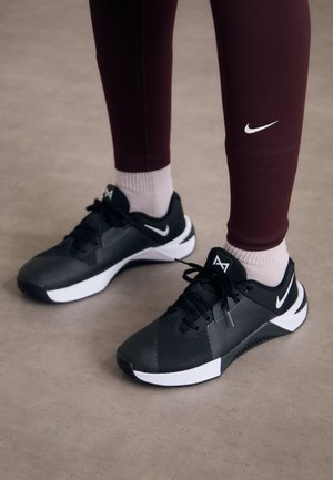 Person iført sorte og hvide Nike-sneakers med hvide sokker og mørkerøde Nike-leggings, der står på en grå gulv.
