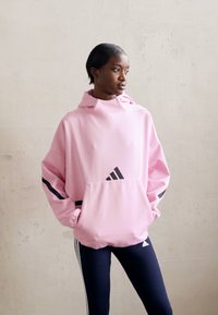 Rožinė per didelė megzta striukė, pagaminta iš minkštos medžiagos, su didele priekinė kišene, su juodomis juostelėmis ir "adidas" logotipu priekyje.
