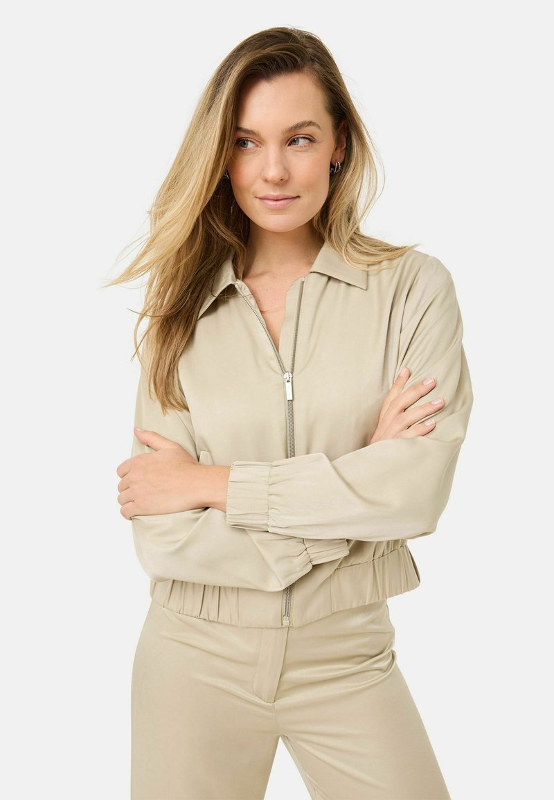 Beige zip-up jacka med krage, elastiska ärmslut och cropped längd. Slätt, glänsande tyg med en subtil lyster.
