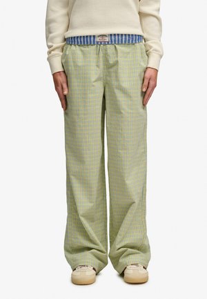 Persoon draagt losse gele en blauwe geruite broek met zakken, witte sneakers en een crèmekleurige trui met zichtbare gestreepte tailleband.