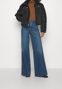 Osoba w czarnej kurtce puchowej, brązowym swetrze z golfem, szerokich niebieskich dżinsach i czarnych butach na tle białego tła.