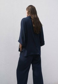 Una mujer con cabello largo se encuentra de pie frente a una pared blanca, vistiendo una camisa azul marino holgada y pantalones a juego, con las manos en los bolsillos.