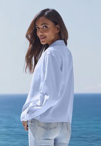 White long-sleeve shirt with a collared neck, button cuffs, and relaxed fit, paired with light denim jeans against a sea backdrop.