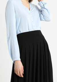 Blusa azzurra chiara a maniche lunghe con scollo a V e chiusure a bottone, abbinata a una gonna nera plissettata. Texture del tessuto liscia.