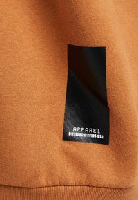 La tela de algodón naranja presenta una etiqueta rectangular negra con el texto "APPAREL" en blanco, creando un contraste en el diseño y la textura.