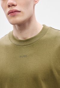 Olivno zelena majica z okroglim izrezom, izdelana iz gladke tkanine z nežno teksturo, na sprednji strani pa ima vtisnjen logotip "HUGO".