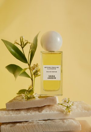 Flacon de parfum en verre transparent avec un bouchon rond blanc. Liquide jaune à l'intérieur. Entouré de feuilles vertes et de petites fleurs blanches sur une pierre texturée.