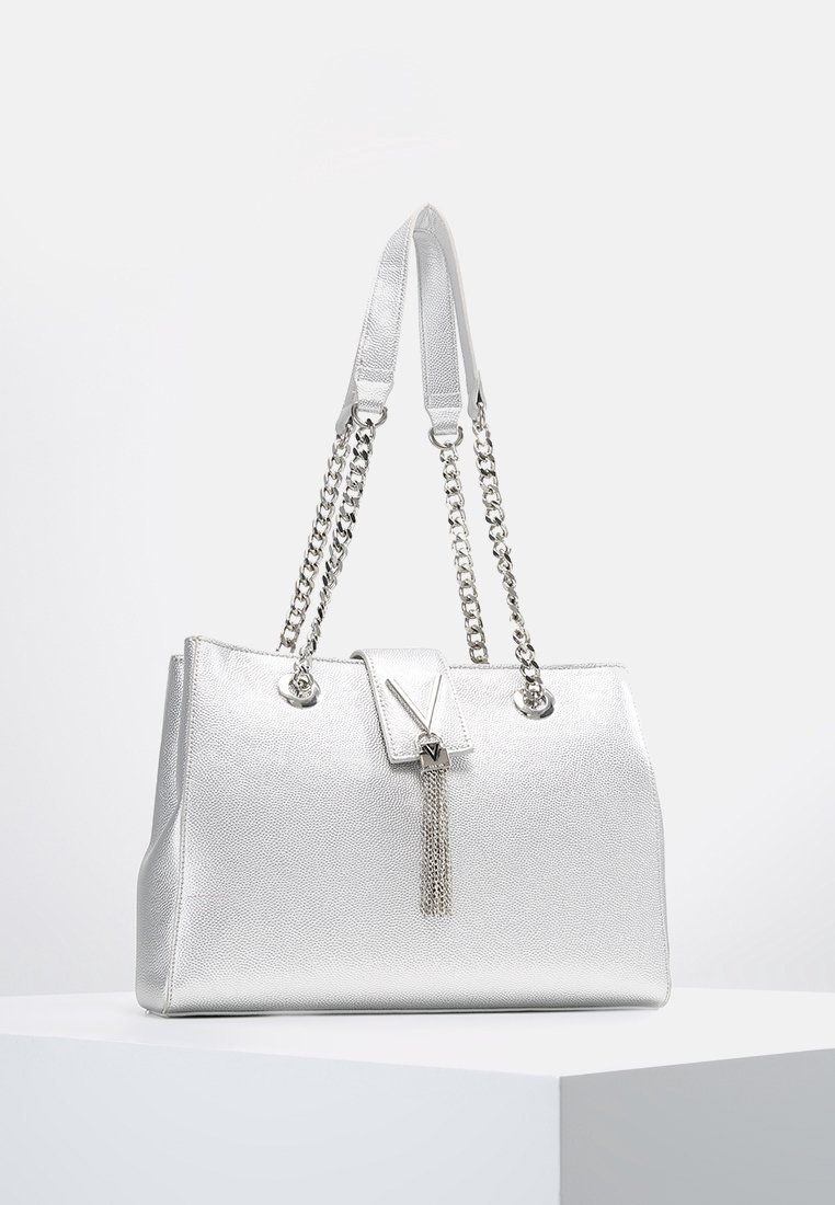 Silverfärgad handväska med strukturerad yta, dubbla kedje- och remhandtag, V-formad lås samt en tofsdetalj fram.