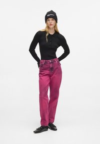 Crna rebrasta majica dugih rukava, ružičaste izblijedjele traperice i crni zimski šešir s bijelim tekstom. Model stoji s rukama na bokovima protiv bijele pozadine.