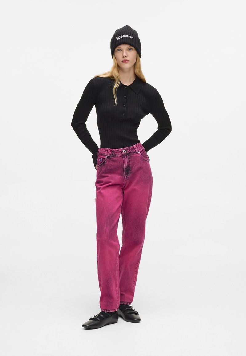 Crna rebrasta majica dugih rukava, ružičaste izblijedjele traperice i crni zimski šešir s bijelim tekstom. Model stoji s rukama na bokovima protiv bijele pozadine.