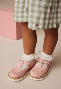 Chaussures en cuir rose avec un design à bride en T, perforations décoratives, semelle blanche, associées à des chaussettes blanches à volants. Robe à carreaux verts visible.