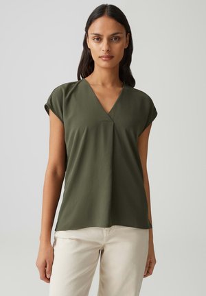 Donna con capelli lunghi e scuri che indossa una camicetta verde oliva a maniche corte con scollo a V e pantaloni beige chiaro su uno sfondo semplice.