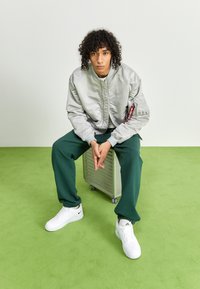 Sølv bomberjakke, grønne joggebukser og hvite sneakers med svarte detaljer. Modell sitter på en grå spinnerkoffert mot et grønt gulv.