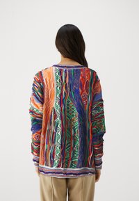 Sweater surdimensionné coloré présentant des motifs vifs en orange, vert, bleu et rouge, avec un tissu texturé et des poignets rayés contrastants.