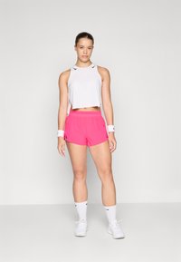 Weißes, kurzes sportliches Tanktop, knallpinke Shorts und weiße Sportschuhe mit weißen Socken. Auffällige schwarze Akzente auf dem Tanktop.