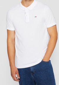 Vit kortärmad polo skjorta i bomull, med en klassisk krage och en liten broderad logotyp på bröstet. Kombineras med blå jeans.