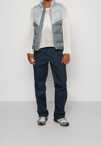 Person klädd i en vit meshtröja, en ljusgrå Nike-padded väst, marinblå byxor och vit-grå sneakers stående mot en neutral bakgrund.