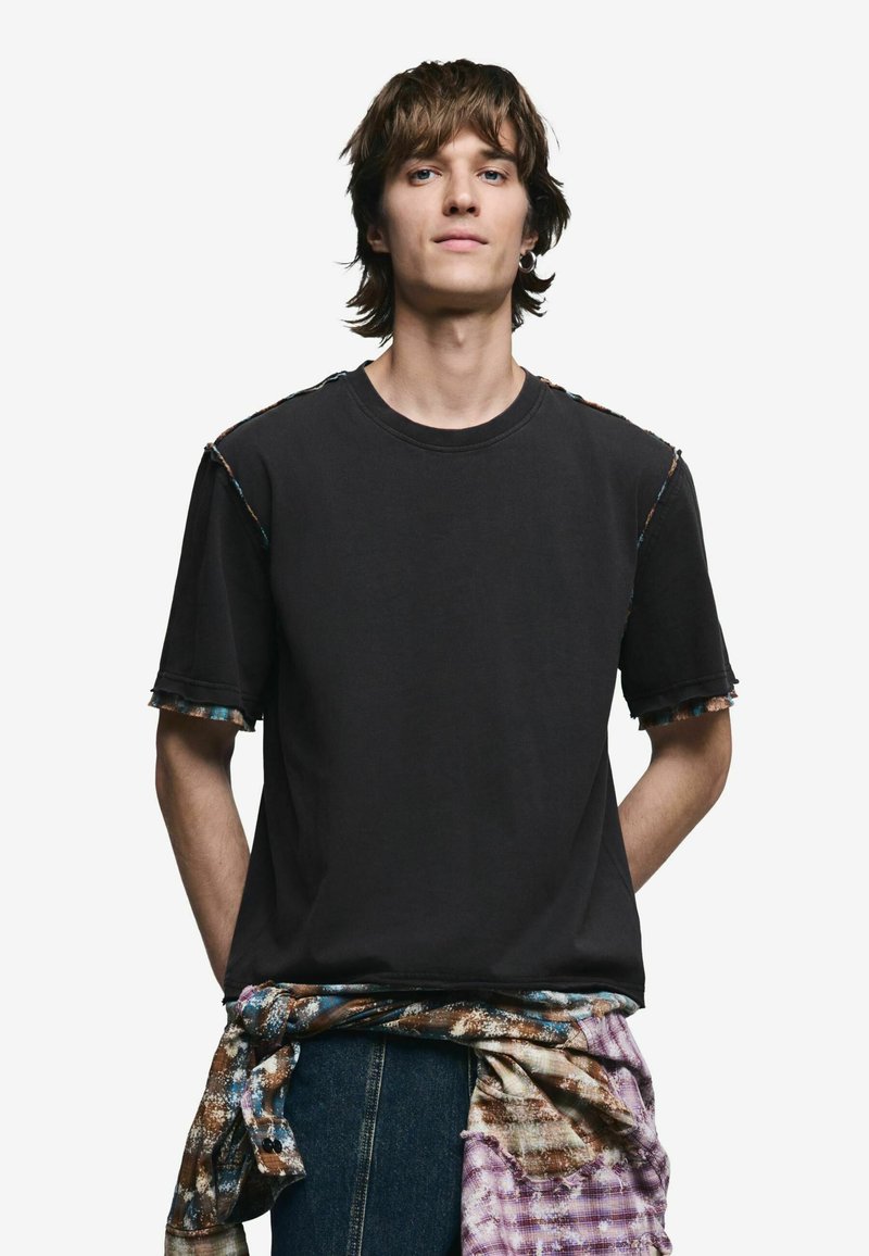 T-shirt noir avec des détails à bords bruts et une bordure colorée le long des épaules, associé à une chemise en flanelle à motifs nouée autour de la taille.