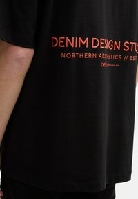 Black cotton t-shirt with short sleeves, featuring red text: "DENIM DESIGN STUDIO NORTHERN AESTHETICS // EST." on the back.