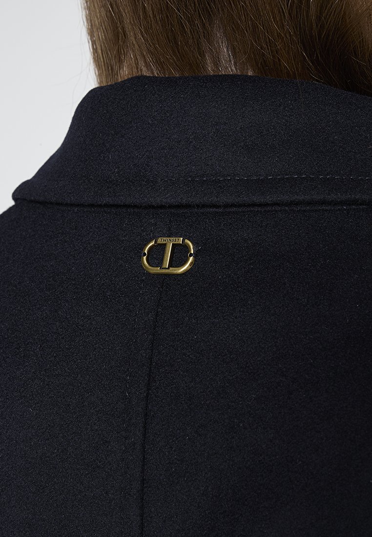Abrigo de lana negro con cuello, con un acento dorado en la parte posterior y un detalle de logo, que enfatiza líneas limpias y un acabado texturizado.