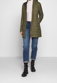 Veste matelassée vert olive avec capuche, dotée d'une fermeture éclair et d'une texture côtelée. Portée sur un pull col roulé blanc et un jean bleu. Bottes noires.