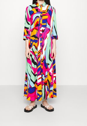 Femme portant une longue robe colorée à motif abstrait avec des boutons sur le devant et des manches trois quarts, associée à des sandales blanches.