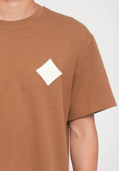Brown short-sleeve t-shirt made of textured fabric, featuring a white diamond-shaped logo patch on the left chest area.