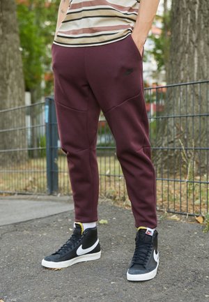 Person iført bordeaux Nike joggingbukser og sorte Nike højtop-sneakers står på en brolagt sti nær et metalhegn og træer.