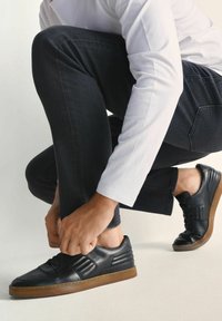 Osoba w białej koszuli z długim rękawem i czarnych dżinsach, którą widać w pozycji klęczącej, wiąże czarne trampki z brązowymi podeszwami na jasnym tle.