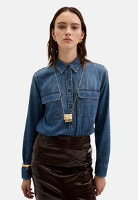 Denim skjorte med knaplukning, to brystlommer, lange ærmer. Sammen med en blank, brun skindnederdel med krokodillemønster og guld halskæde.