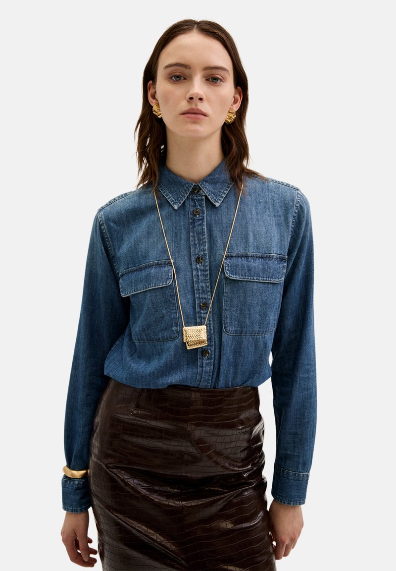 Denim skjorte med knaplukning, to brystlommer, lange ærmer. Sammen med en blank, brun skindnederdel med krokodillemønster og guld halskæde.