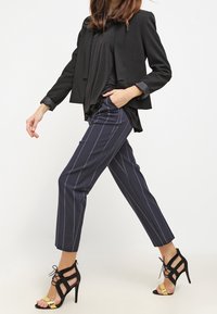 Blazer nero, top nero fluido, pantaloni navy a righe e décolleté neri con cinturini e dettagli dorati. Modella che cammina su uno sfondo neutro.