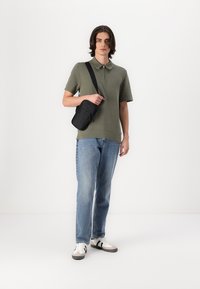 Grünes kurzärmliges Poloshirt mit einem Reißverschlusskragen, hellblaue Jeans aus Denim und weiße Sneaker mit schwarzen Akzenten; hält eine kleine schwarze Umhängetasche.