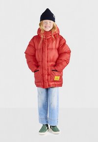 Rød dunjakke med hætte, med sorte detaljer og en gul label. Båret over lyseblå jeans og grønne sko med prikker.