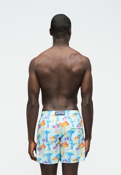 Άνδρας στέκεται με την πλάτη προς την κάμερα, φορώντας πολύχρωμα μαγιό με σχέδιο μέδουσας, σε απλό φωτεινό φόντο.