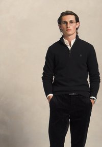 Tânăr cu ochelari, îmbrăcat cu un pulover negru cu fermoar peste o cămașă albă cu guler, cu mâinile în buzunarele pantalonilor negri, pe un fundal simplu.
