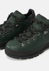 Πράσινα δερμάτινα παπούτσια πεζοπορίας με μαύρες ελαστικές σόλες, με μεταλλικά άγκιστρα και κορδόνια, και διάτρητες λεπτομέρειες στο γιακά.