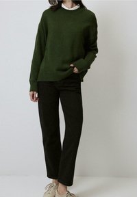 Personne portant un pull vert foncé, un pantalon noir à jambes droites et des baskets beige clair sur un fond clair uni.