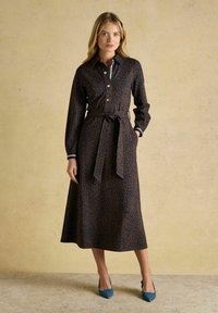 Sieviete stāv garās piedurknēs, brūnā rakstainā midi kleitā ar sasietu jostu, apvienojot ar ziliem norādītiem papēžiem.